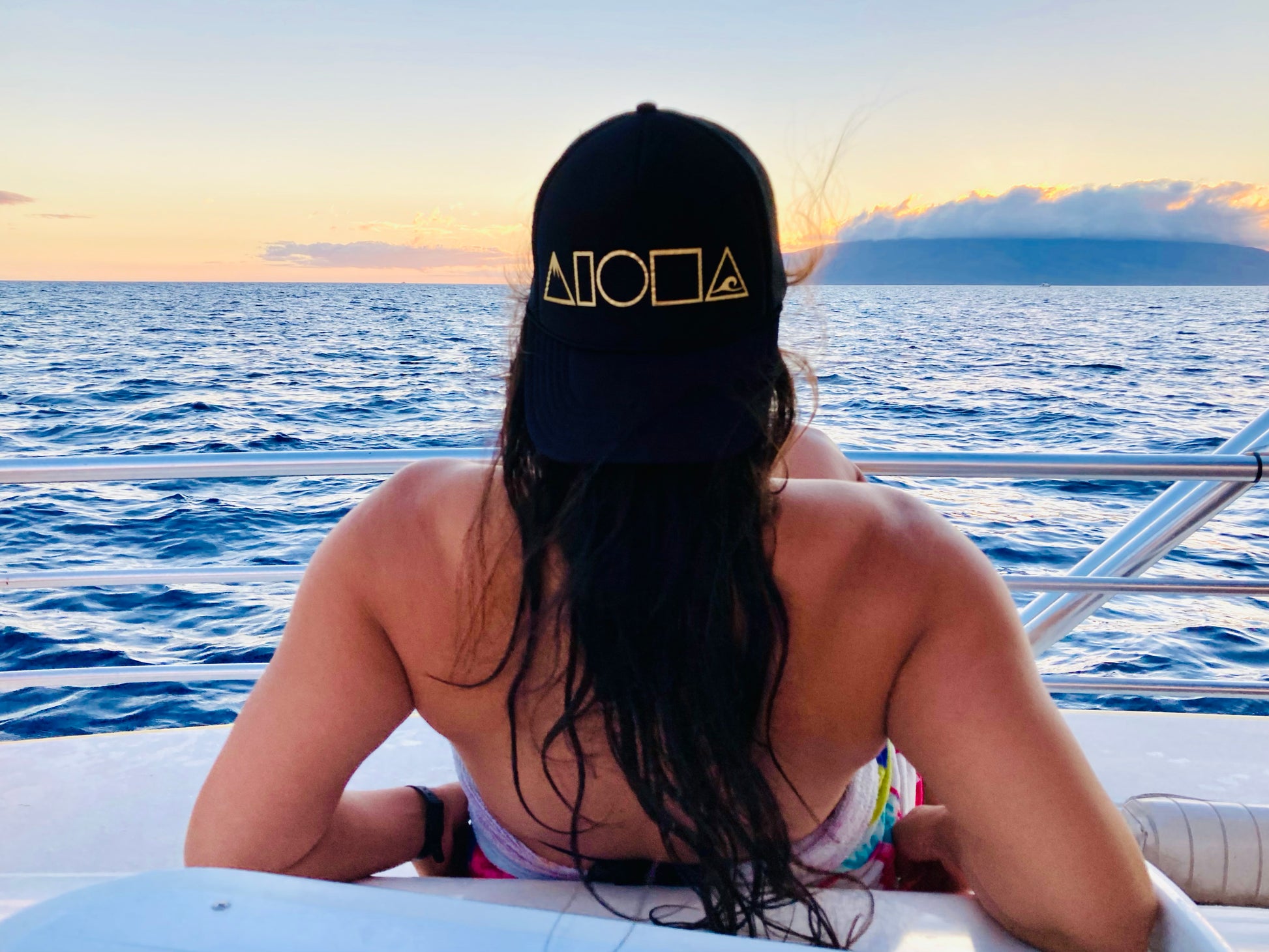 Person on a boat in Maui Hawaii watching the sunset with a Mauka to Makai trucker hat turned backwards